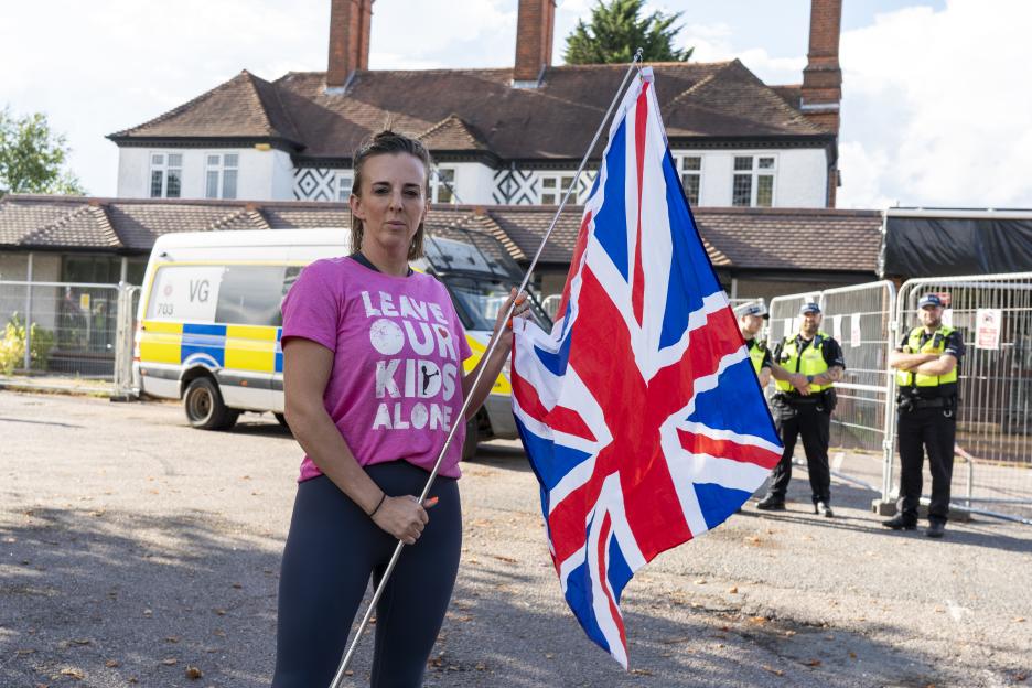 Woman holding a Union Jack flag and wearing a "Leave Our Kids Alone" shirt, with police officers in the background.