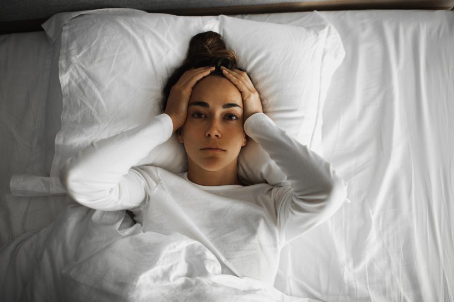 Woman lying in bed, hands on head, looking distressed.