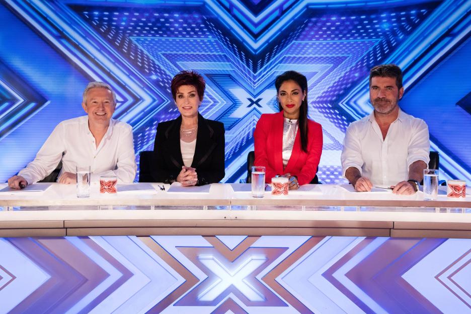 X Factor judges Louis Walsh, Sharon Osbourne, Nicole Scherzinger, and Simon Cowell sitting at a desk with blue 'X Factor' backdrop.