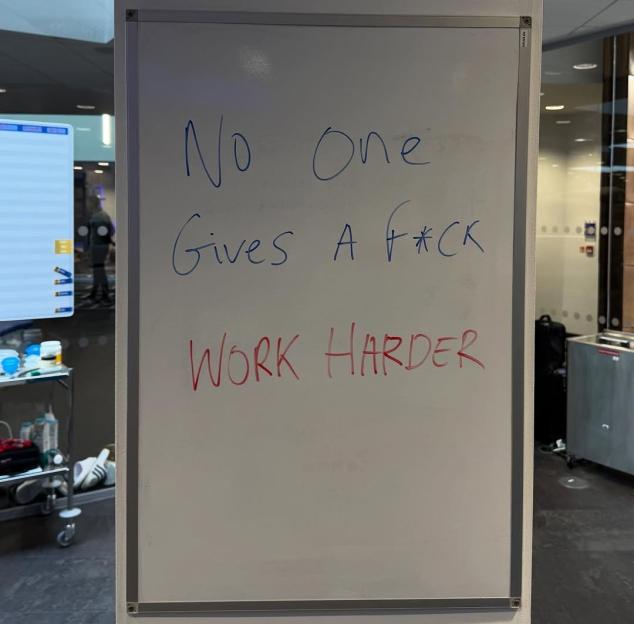 A whiteboard with "No One Gives A F*CK" in blue and "WORK HARDER" in red.