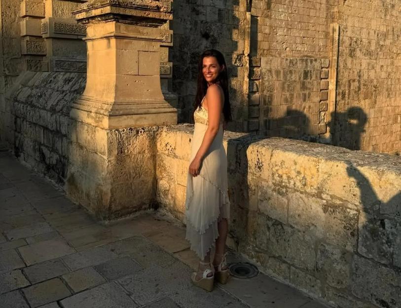 Marisa Abela in a light-colored dress, standing near an ancient stone wall.