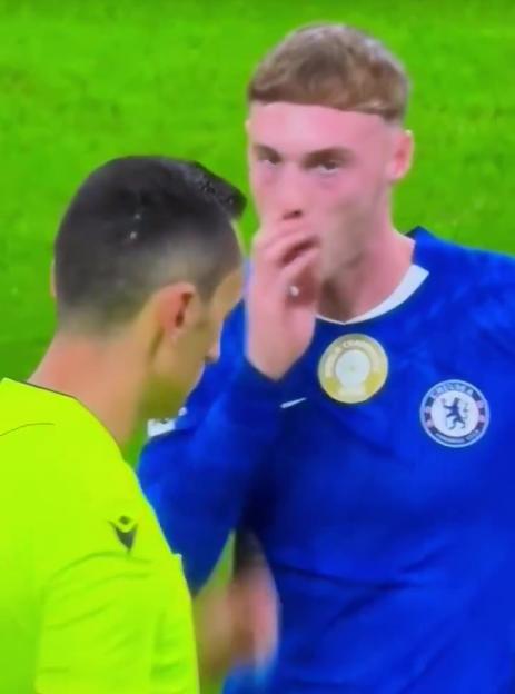 Cole Palmer of Chelsea FC in a blue jersey, with a hand to his mouth, facing a referee in a neon yellow shirt.