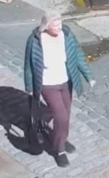 A light-skinned person with light hair wearing a dark blue coat, light shirt, and dark pants walking on a cobblestone surface.