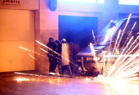 Riot police with shields facing a van amidst exploding fireworks.