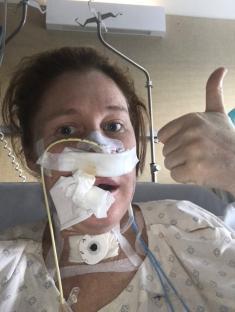 Woman in hospital bed giving thumbs up, post-facial surgery.