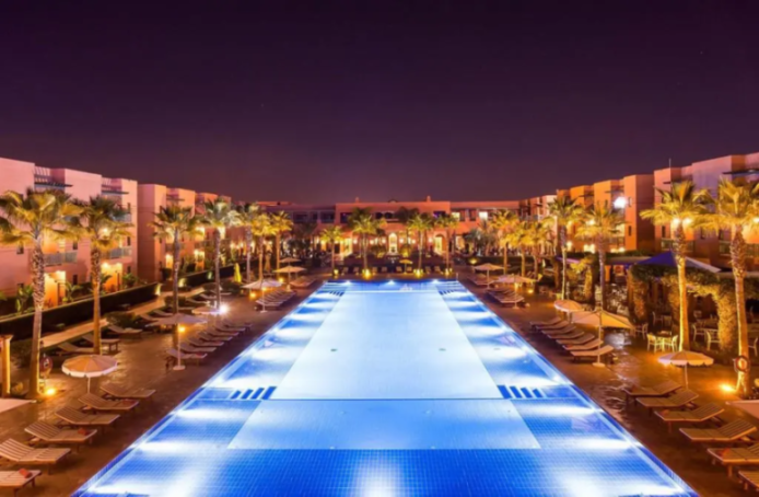 Nighttime view of JAAL Riad Resort in Marrakech, Morocco, with a large, illuminated swimming pool, palm trees, and resort buildings.