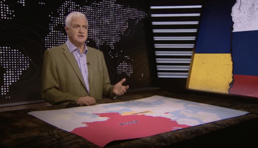 A man in a tan jacket standing behind a table with a map of Russia and Ukraine, with a background of a world map and a Russian-Ukrainian flag.
