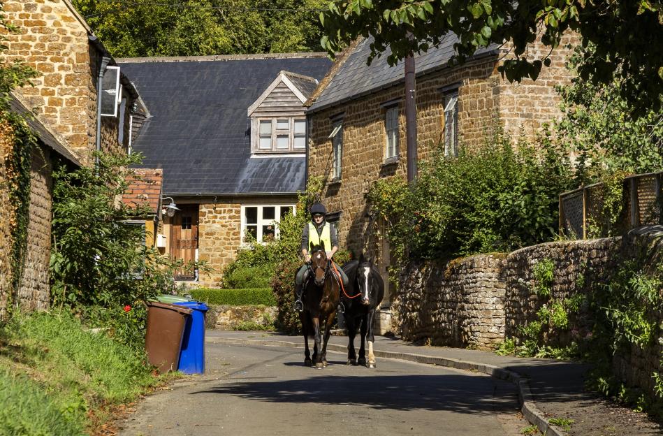 Person riding two horses down a narrow street in a Cotswolds village.