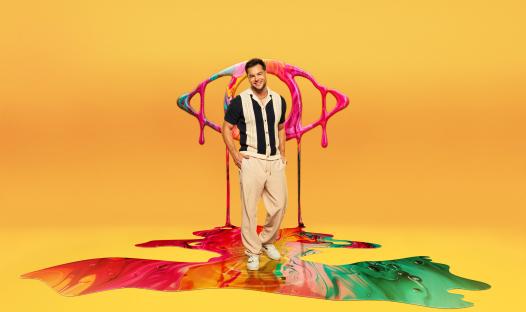 Chris Hughes standing on a colorful paint spill, in front of a paint-splattered eye graphic.