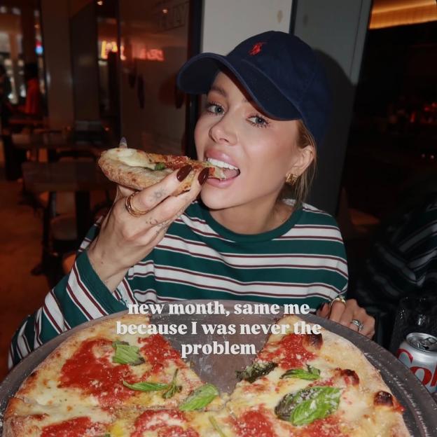 Woman in a baseball cap eating a slice of pizza.