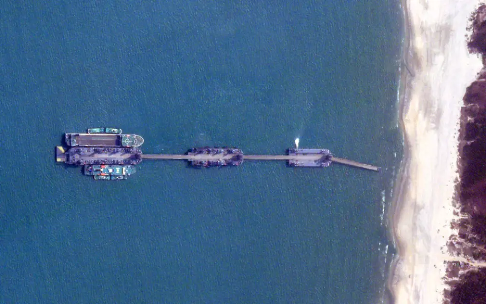 Aerial view of a pier connecting to the shore with multiple boats docked, some with structures, and a sandy beach to the right.