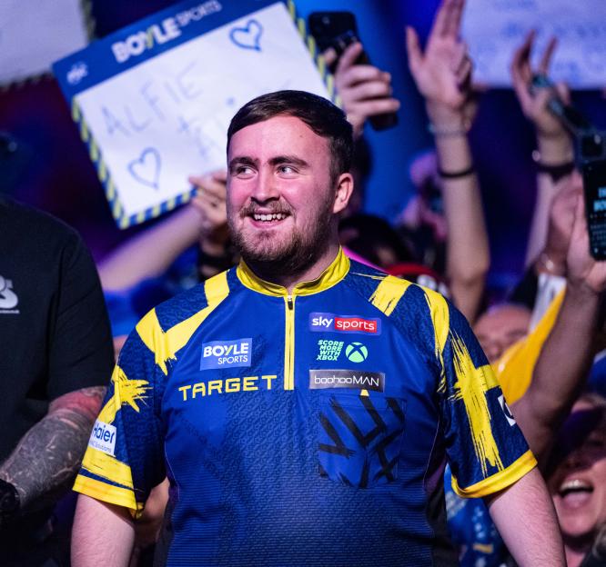 Luke Littler smiling at the crowd while wearing a blue and yellow jersey.