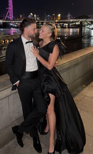 Jojo Siwa in a black thigh-split dress with Chris Hughes at night with a city skyline in the background.