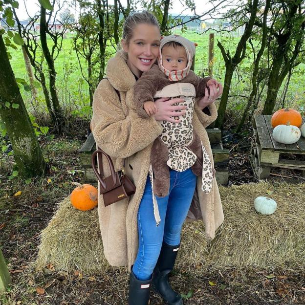 A woman in a fur coat and jeans holding a baby in a leopard print carrier at a pumpkin patch.