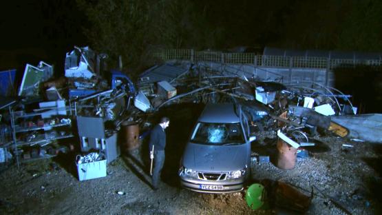 A car with a shattered windshield sits amidst debris from a damaged structure at night.