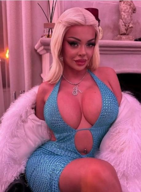 A woman with blonde hair, wearing a sparkly blue dress, a fur coat, and a "K" pendant.