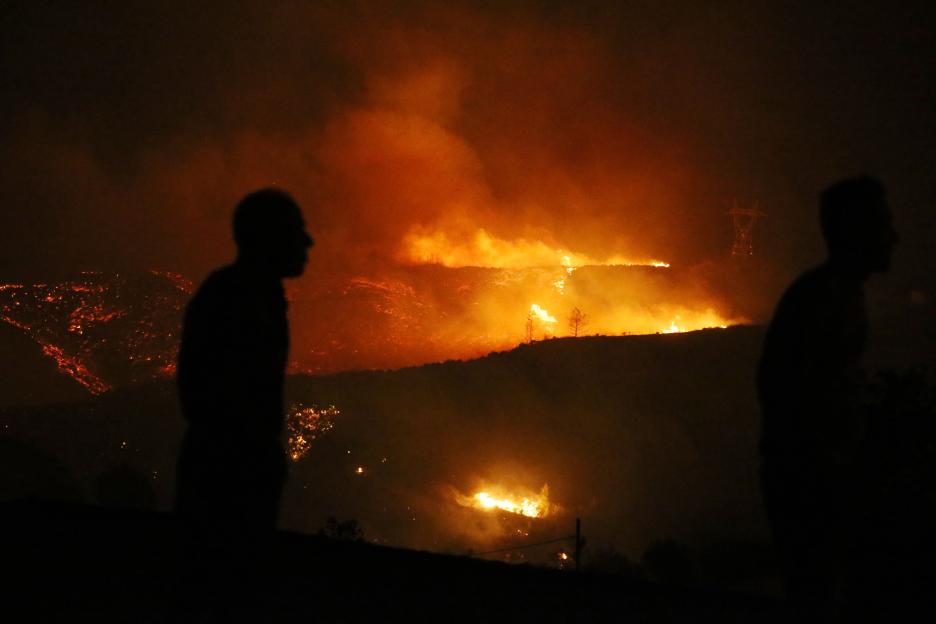 Silhouettes of two people observe a large wildfire burning across a hillside at night.