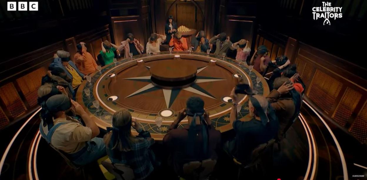 Contestants on The Celebrity Traitors with blindfolds covering their eyes, seated around a large circular table.