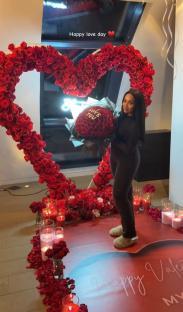 Woman standing in front of a heart-shaped rose arrangement holding a bouquet of roses that says