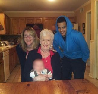 Photo of a family in a kitchen; a woman holds a baby, flanked by another woman and a young man.