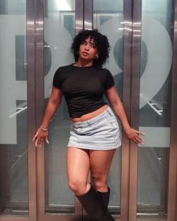 Woman in black shirt and denim mini skirt leaning against elevator doors.