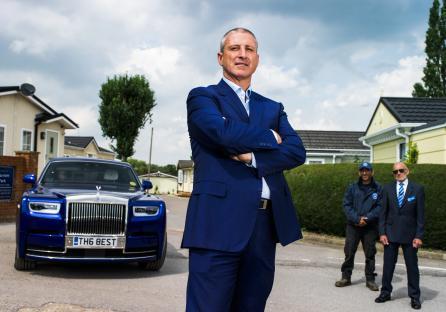 A man in a suit stands in front of a blue Rolls Royce in a residential park. Two security guards stand behind him.