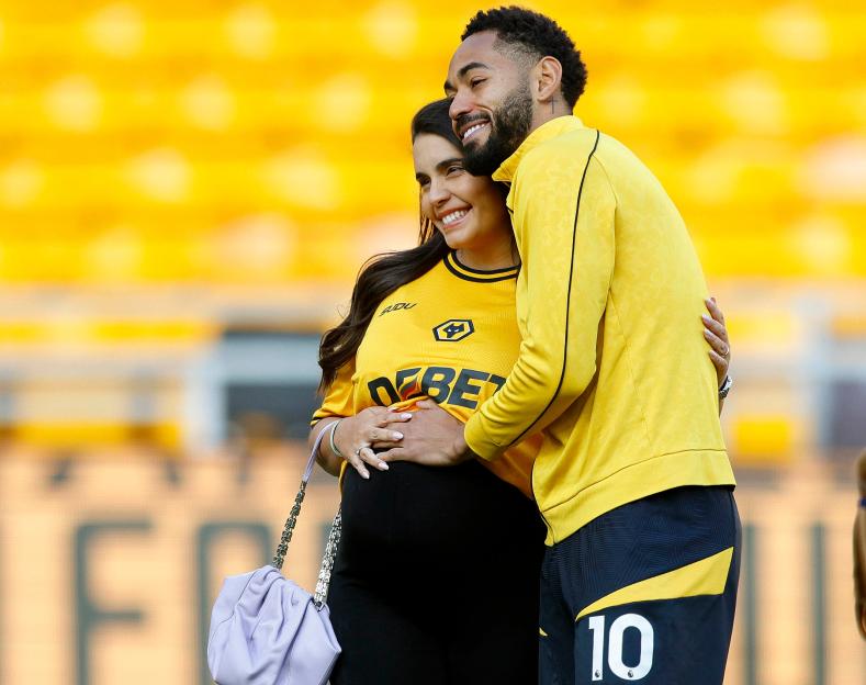 Matheus Cunha of Wolverhampton Wanderers and his pregnant wife embracing on a soccer field.