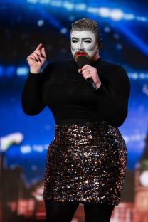 Person in black top and sequined skirt with mime makeup holding a microphone.