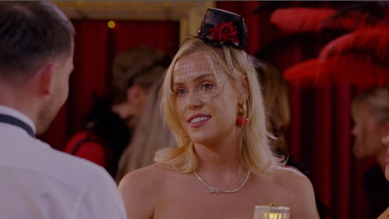 A blonde woman wearing a small hat and veil speaks to a man.