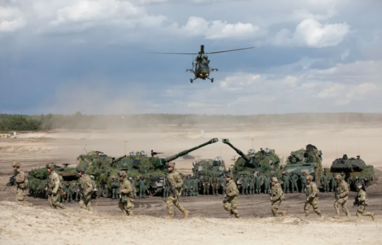 Military exercise with a helicopter flying over tanks and soldiers on a dusty field.