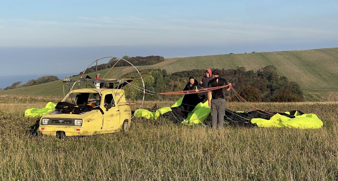 A yellow three-wheeled car with a paramotor frame on top in a grassy field with people preparing a parachute.
