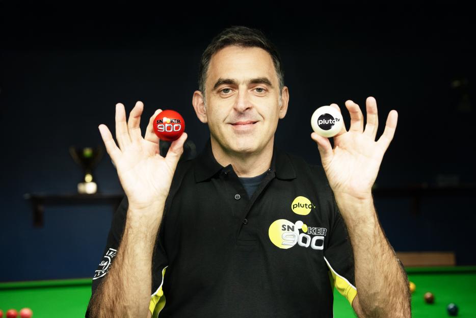A man in a black polo shirt holding a red snooker ball and a white Pluto TV ball.