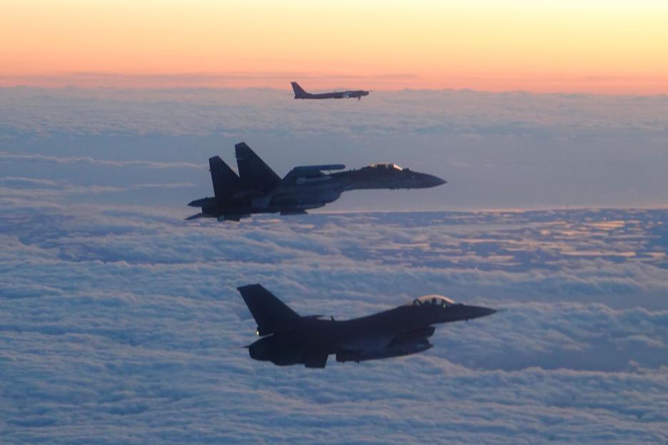 An F-16 fighter jet in the foreground escorts a Russian Su-35 fighter, with a Tu-95 bomber in the distance, flying above clouds at sunset.
