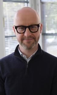 Portrait of a bald man wearing glasses and a dark sweater.