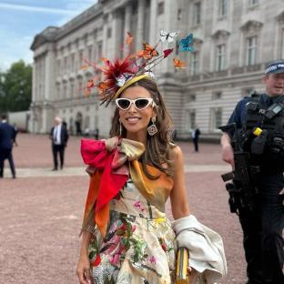 Woman in floral dress and butterfly hat outside Buckingham Palace.