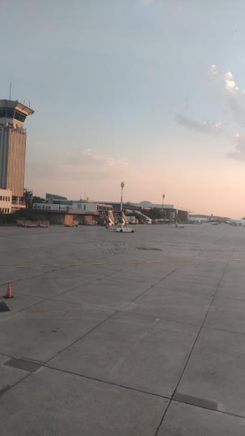 Airport tarmac with a control tower, buildings, and planes under a light blue and pink sky.