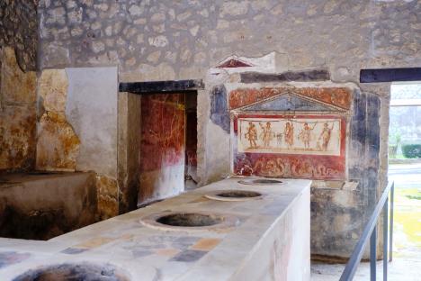Ancient Thermopolium with Roman Fresco, it is House and Thermopolium of Vetutius Placidus, The thermopolium of Vetutius Placidus opens on via dell’Abbondanza and represents social mobility in Pompeii in Roman times, where merchants and craftsmen also held a high social status, reserved only to landowners in older times. Drinks and hot food were served in this place, as the name indicates, stored in large jars placed in the richly decorated masonry counter of the tavern. photo taken in a street in Pompei called