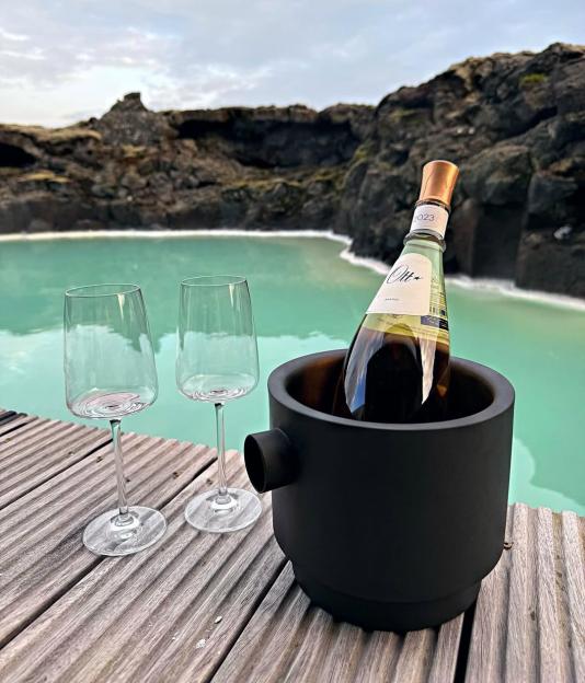 Wine bottle in a cooler with two wine glasses on a wooden deck overlooking a geothermal pool.