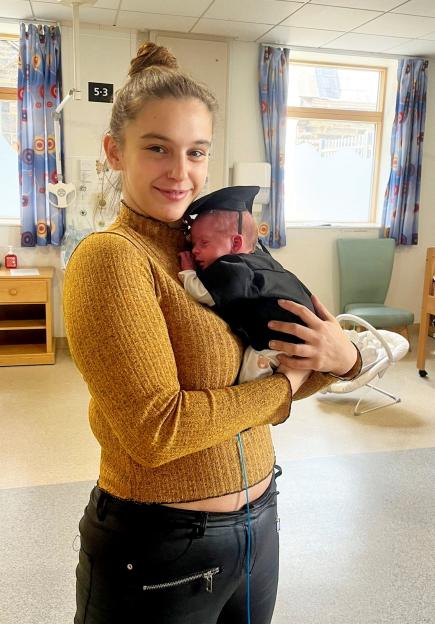 Megan Isherwood holding her newborn son, Jackson, who is wearing a tiny graduation cap and gown in a hospital room.