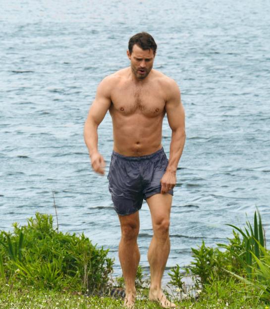 Jamie Dornan in wet swim trunks by a lake.