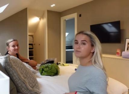 Two women in a bedroom; one sits on a bed, the other stands.