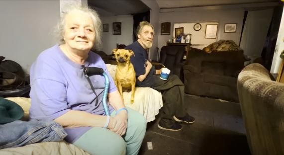 A photo of two elderly people and a small dog sitting on a couch in a cluttered home.