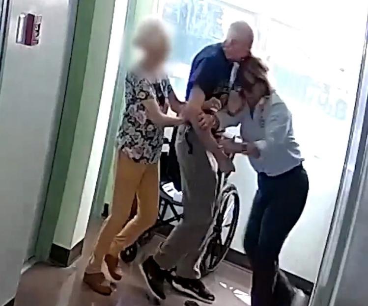 Video still of three people struggling around a wheelchair in a hallway.