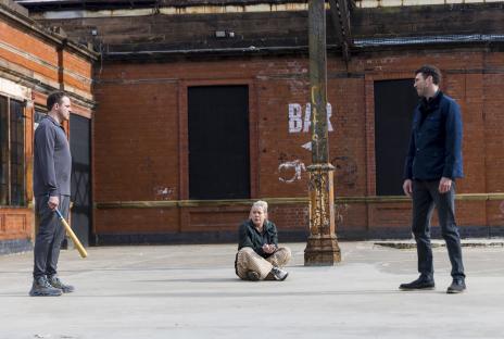 Scene from Coronation Street: a woman sits on the ground between two men, one holding a baseball bat.