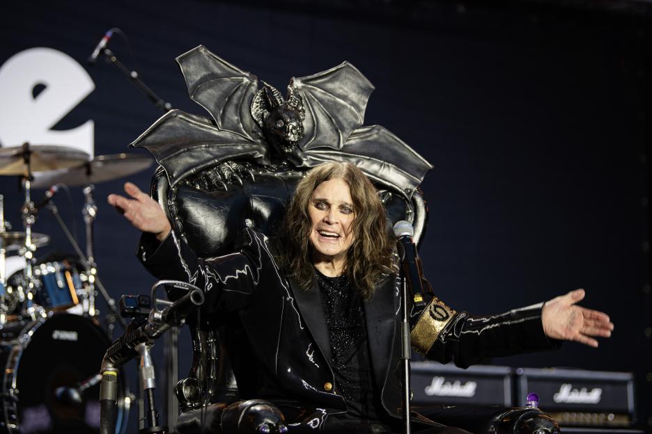 Ozzy Osbourne sitting in a black leather throne with a bat-like headrest, speaking into a microphone.