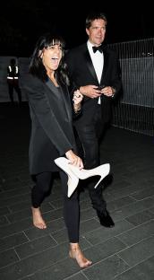 A woman in a black blazer and leggings walks barefoot, holding her white high heels, alongside a man in a tuxedo.