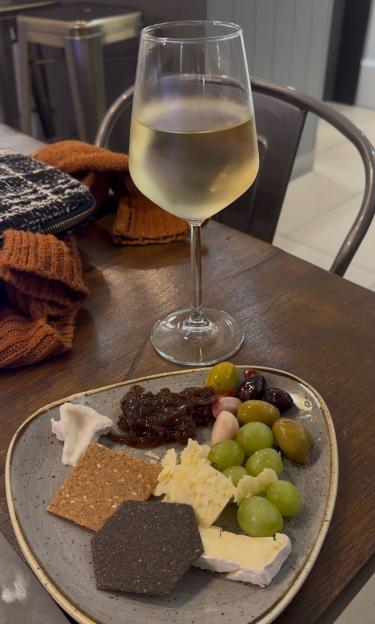 A glass of white wine and a plate of cheese, crackers, grapes, olives, and preserves on a wooden table.