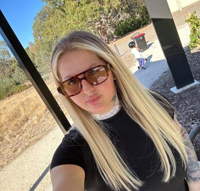 Woman wearing sunglasses and a neck brace, with a child visible in the background.