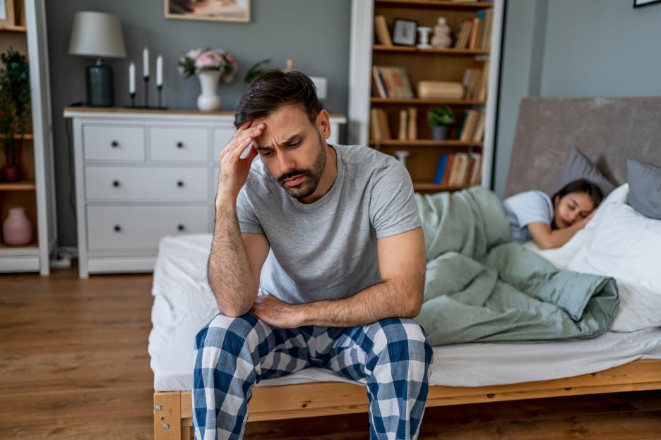 Man sits on bed looking distressed while his wife sleeps.