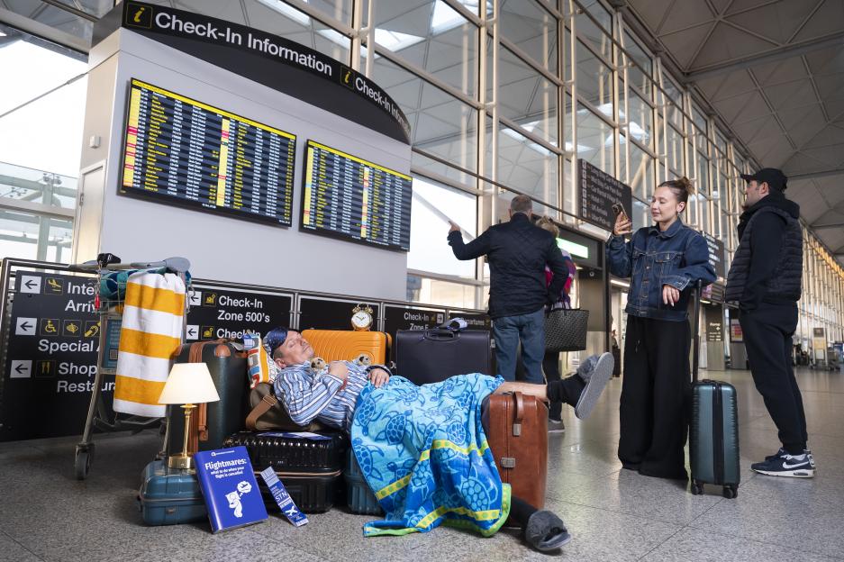 Man asleep on luggage at an airport check-in, wearing pajamas and covered by a blanket, next to flight information screens and other waiting passengers.